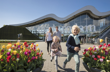 Een vrolijk gezin met twee kinderen loopt tussen kleurrijke tulpen richting de ingang van Madurodam, met een modern gebouw met golvend dak op de achtergrond onder een heldere blauwe lucht.