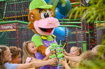Een vrolijke mascotte in de vorm van een aap deelt bekers uit aan lachende kinderen in een kleurrijke indoor speeltuin.