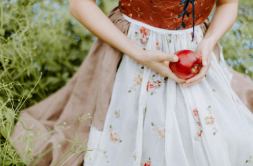 Vrouw in een sprookjesachtige jurk met bloemenmotief houdt een rode appel vast, omringd door groen gras en planten.
