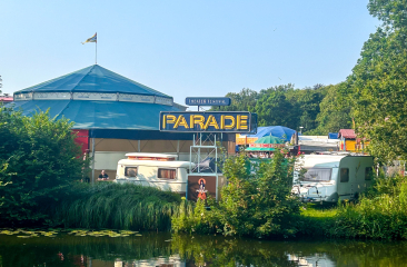 Circustenten en kleurrijke wagens van festival De Parade staan opgesteld in een groen parklandschap, weerspiegeld in het rustige water van een vijver onder een heldere blauwe lucht.