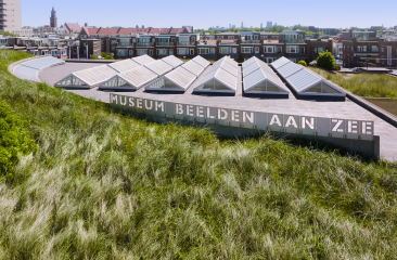 Luchtfoto van Museum Beelden aan Zee in Scheveningen, met een grasduin op het dak en de skyline van Den Haag op de achtergrond.