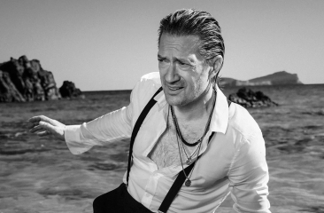 Zwart wit foto van Joost Spijkers die uit de zee komt lopen. Hij staat op een tropisch strand met rotsen en draagt een zwarte broek met bretels en een witte blouse. Promotiebeeld voor zijn show: De broer van Aznavour. 