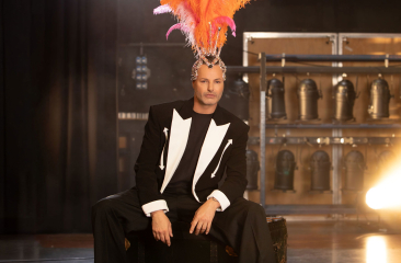 Fred van Leer zit op de rand van het podium met een zwart/wit pak aan en een showgirl hoofdtooi met oranje/roze veren op zijn hoofd. Hij kijkt serieus de verte in. 