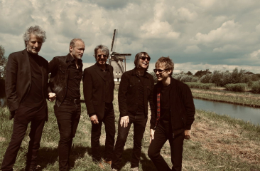 De leden van coverband Timebox poseren lachend in zwarte outfits op een dijk met een molen op de achtergrond.