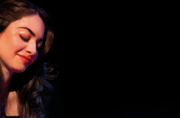 Close-up of Muza Valiaddinzade while playing the piano in front of a dark backround.. She is looking down at her hands. 