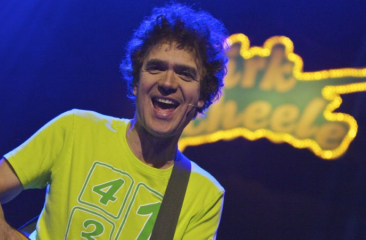 Dirk Scheele vrolijk en spelend op gitaar op podium in geel shirt en geel bandlogo met zijn naam erin passend bij zijn kindervoorstelling popconcert Foto: Guido Breedveld