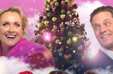 Joke de Kruijf staat samen met Winston Post lachend voor een versierde kerstboom. De achtergrond bestaat uit een roze wolkenlucht en een vliegtuig. 