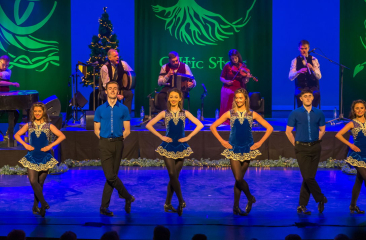 De leden van Celtic Steps staan op een rij op het podium in blauwe danskostuums. Achter hen staan 6 muzikanten. 