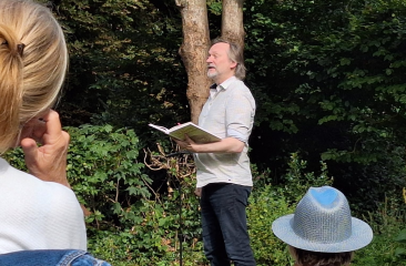 Man leest voor uit een boek tijdens een buitenoptreden in een groene, bosrijke omgeving; toeschouwers luisteren vanaf stoelen op het gras.
