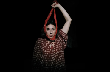 Promotiebeeld dansclick Melisa Diktaş in een rode bloemenjurk kijkt recht in de camera, met een rode stoffen band strak rond haar gezicht en omhooggehouden arm, tegen een donkere achtergrond.