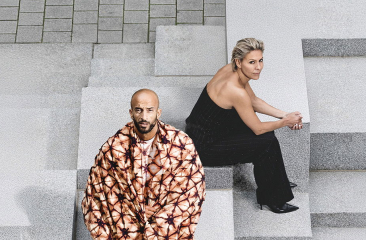 Foto van Mouna en Walid van schuin bovenaf. Ze zitten op grijze betonnen blokken en kijken recht in de camera. Mouna draagt een zwarte jumpsuit en khalid een rood-oranje tie-dye jas. 