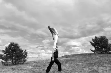 Twee mensen doen een acrobatische lift op een open veld met bomen en een bewolkte lucht 
