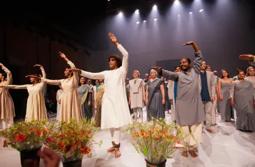 Groep dansers op een verlicht podium maakt een buiging, gekleed in losse, kleurrijke outfits met bloemen op de voorgrond.