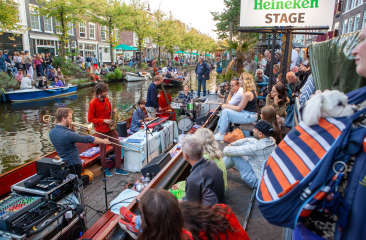 Muzikanten spelen op een boot in een gracht, publiek zit langs de kade bij het Heineken Stage-bord