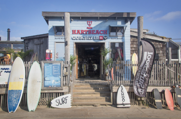 Ingang Hart Beach surfschool bestaande uit lichtblauwe houten panelen en rood logo. Er staan surfplanken op een rij voor de deur naast de trap. Ingang Hart Beach surfschool bestaande uit lichtblauwe houten panelen en rood logo. Er staan surfplanken op een rij voor de deur naast de trap.
