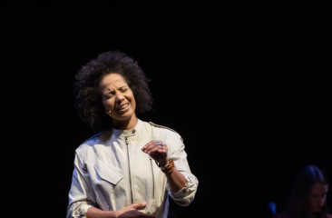 Vrouw in wit kostuum legt emotioneel iets uit op donker podium