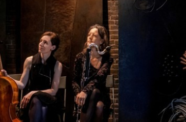Katharina Gross en Fie Schouten zitten naast elkaar op het podium met hun instrumenten: de cello en de basklarinet. Ze dragen donkere kleding en kijken geamuseerd naar de linkerkant. 