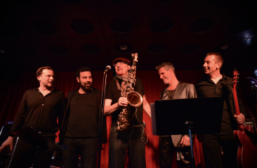 Bandfoto met de bandleden in donkere achtergrond met rood doek en een spot op hun gericht passend bij hun Bowie interpretatie.