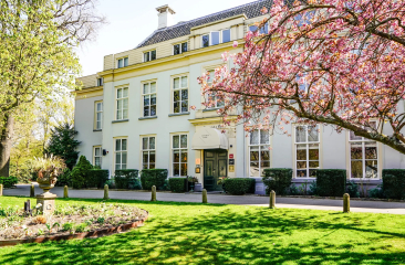 Wit klassiek gebouw in Den Haag met bloeiende boom, groene tuin en zonlicht op de voorgevel Wit klassiek gebouw in Den Haag met bloeiende boom, groene tuin en zonlicht op de voorgevel