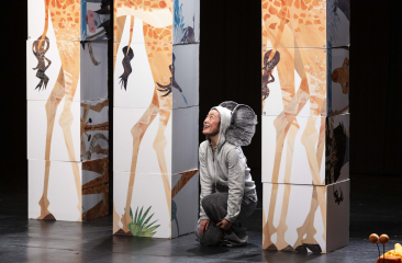 Een vrouw verkleed als olifant zit op een podium omringt door decorstukken die giraffen voorstellen. 