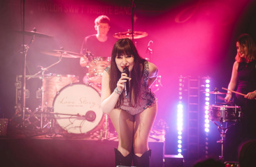 De leden van de Taylor Swift Tribute Band staan op een roze verlicht podium. De zangeres kijkt lachend de camera in. 