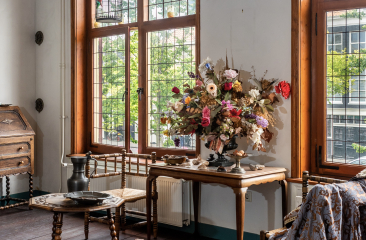 Historisch interieur met houten meubels, droogbloemen op een tafel en uitzicht op grachtenpand door glas-in-loodramen Historisch interieur met houten meubels, droogbloemen op een tafel en uitzicht op grachtenpand door glas-in-loodramen