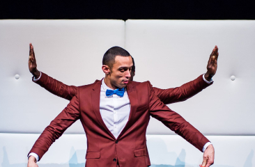 Man in rood pak met blauwe strik op podium, met extra armen zichtbaar door tweede persoon achter hem