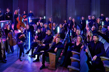 Leden van het WDR Funkhausorchester zitten met hun instrumenten op stoelen in een theaterzaal