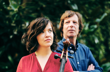 Twee mensen staan buiten voor een groene achtergrond; de vrouw vooraan houdt een cello vast, de man staat erachter.