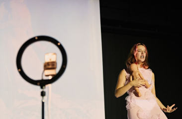 Vrouw in roze outfit performt voor een projectiescherm, met een ringlamp en smartphone op statief op de voorgrond.