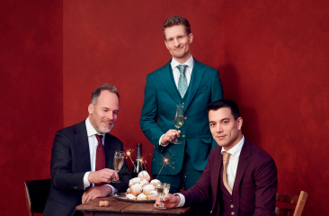Joep Hullegie en Andries Tunru zitten aan een tafel met oliebollen, champagne en sterretjes. Stefan Hendrikx staat aan het hoofd van de tafel. De mannen zijn gekleed in pakken met stropdassen en staan voor een rode achtergrond. 