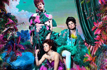 Drie performers van Collectief Aqueerius in extravagante barok-wave kostuums poseren in een kleurrijke, fantasierijke setting. De visuele stijl is een mix van barok en popart