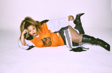 Een vrouw met krullend haar ligt ontspannen op de grond in een studio, draagt een oranje oversized shirt, visnetpanty’s en hoge zwarte laarzen, en kijkt zelfverzekerd in de camera.