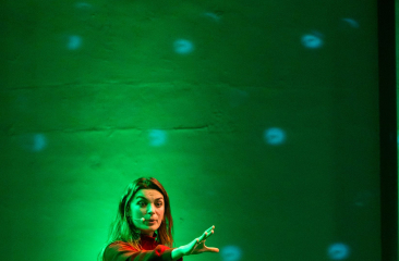 Vrouw op een groen verlichte podiumvloer tijdens een presentatie of theatervoorstelling.