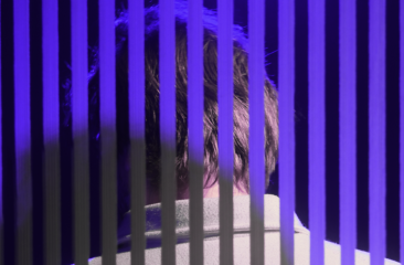 Achteraanzicht van een persoon in paars licht, gefilterd door verticale zwarte strepen die een gevangenis- of blindsuggestie oproepen.