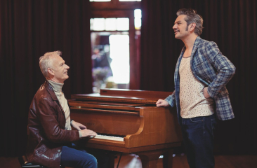 Twee mannen bij een houten piano, één spelend en de ander staand en lachend in gesprek