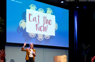 Spreker op podium voor groot scherm met tekst “Eat the Rich”, publiek steekt handen op in zaalsetting