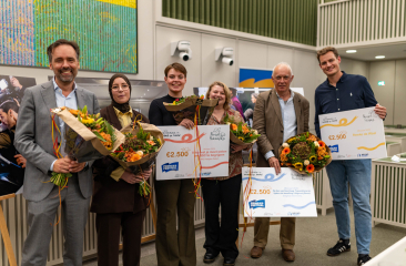 Vijf mensen houden bloemen en cheques tijdens een prijsuitreiking.