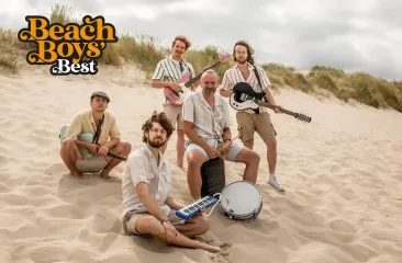 Vijf muzikanten zittend op een zandstrand met instrumenten.