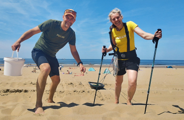 A man and woman stand on the beach, he wears a T-shirt and shorts, she in a yellow shirt and activewear.