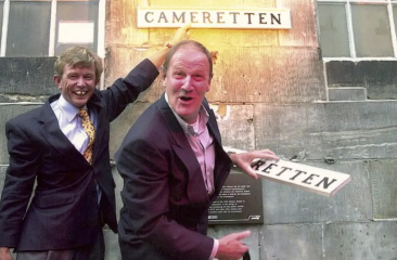 Twee lachende mannen houden een bord met 'RETTEN' vast.