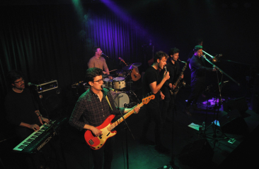 Zes muzikanten spelen live op een podium met instrumenten.
