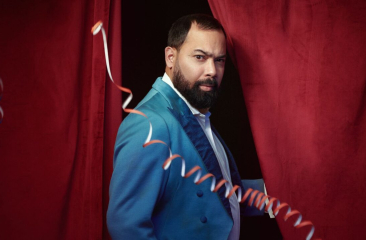 Een man met een baard in een felblauw kostuum staat voor een rode gordijn.