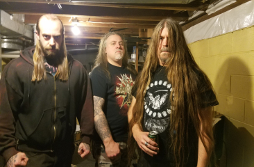 Three members of the band Drawn and Quartered, with long hair and dark clothing, stand in a basement.