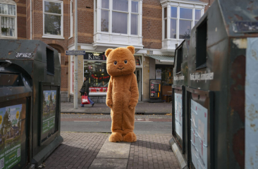 Mascotte in oranje kattenkostuum poseert vrolijk in een theateromgeving, omringd door kinderen en buurtbewoners. De setting ademt een gezellige, laagdrempelige sfeer passend bij een buurtvoorstelling.