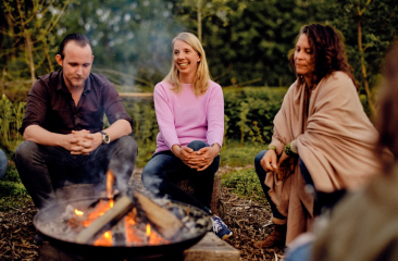 Drei Personen sitzen in der Natur um ein Lagerfeuer.