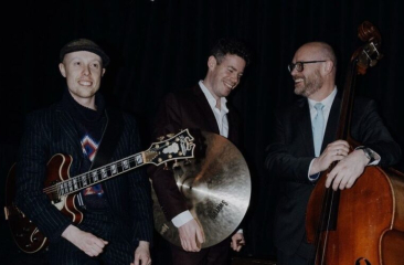 Drie muzikanten met een gitaar, cymbaal en contrabas glimlachen tijdens een optreden.