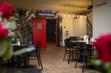 Blick auf den Eingang zu einem Theatersaal mit roter Tür und Tischen.