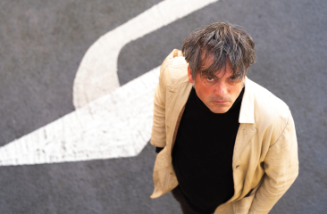 Een man met een lichte jas kijkt omhoog, staand op een straat met een witte pijl.