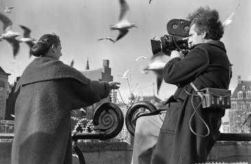 Historische zwart wit foto van een man die met een 8 mm camera een vrouw filmt die meeuwen voert